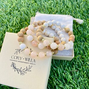 May include: Three aromatherapy bracelets with wooden, white, and pink beads are displayed on green grass. The bracelets are in a beige box with the text "CORY + WILLA HANDMADE AROMATHERAPY JEWELRY". A small drawstring bag is also visible.