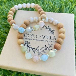 May include: Two beaded bracelets featuring a mix of round wooden beads, white marbled beads, and faceted pastel-colored gemstone beads separated by small silver-toned spacers. The bracelets are resting on a tan box labeled CORY WILLA HANDMADE AROMATHERAPY JEWELRY.