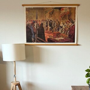 Op de afbeelding: Een vintage poster die een historische scène afbeeldt met een groep mensen in kledij uit die tijd. De poster is ingelijst met een houten rol aan de boven- en onderkant. De poster hangt aan een witte muur met een houten vloerlamp en een houten tafel met een potplant aan de rechterkant van de afbeelding.