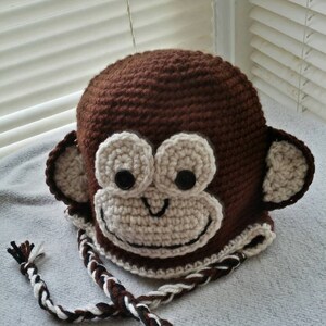 May include: A brown crocheted monkey hat with a white face and black eyes. The hat has a long brown, white, and black braided cord.
