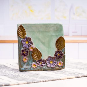 May include: A square ceramic napkin holder with a light green glazed surface. The front features a textured blue base decorated with sculpted purple flowers with yellow centers and brown leaves arranged in a curved pattern along the side.