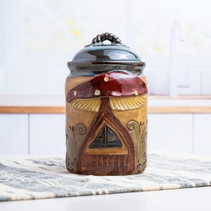 May include: A cylindrical ceramic canister with a dark blue lid featuring a handle. The body is tan with a relief design of a large red mushroom cap over a brown arched door and window, accented by carved swirling vine and floral patterns.