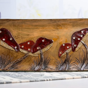 May include: A rectangular ceramic tray with a brown, textured background featuring a raised, hand-painted relief of four red mushrooms with white spots. The mushrooms are depicted growing from dark brown grass, with a glossy finish on the caps.
