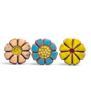 May include: Three ceramic flower-shaped buttons arranged in a row. The first button is peach with a yellow spiral center. The second is blue with a yellow dotted center. The third is yellow with a small red center. Each has eight petals.