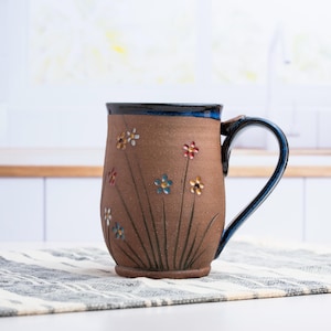 May include: A brown ceramic mug with a dark blue rim and handle. The exterior features hand-painted floral motifs with small red, blue, and white flowers on thin green stems that extend from the base of the mug toward the top rim.