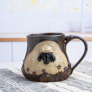 May include: A dark brown ceramic mug featuring a raised, textured cream-colored sheep with a black face and ears. The sheep is surrounded by small purple flowers and brown grass at the base. The mug has a curved handle and a glossy finish.