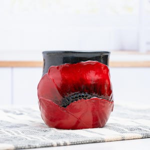 May include: A ceramic mug featuring a glossy black rim and a vibrant, textured red poppy flower design that wraps around the exterior. The petals are sculpted with raised details and deep black centers, creating a bold contrast against the dark background.