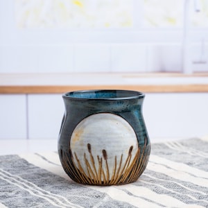 May include: A ceramic mug featuring a dark blue and teal glaze with a prominent circular white moon design. The lower portion of the mug displays a carved pattern of tall, thin brown reeds or cattails extending upward from the base.
