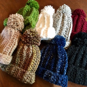 May include: A collection of eight knitted hats with pom-poms in various colours, including beige, green, white, grey, red, brown, blue, and black.