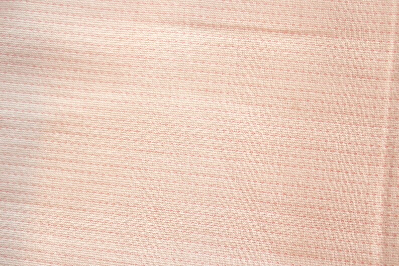 May include: Close-up of a light pink fabric with a textured weave. The fabric has horizontal lines of stitching and a subtle, dotted pattern, creating a soft, tactile appearance. The overall effect is a delicate, neutral textile.