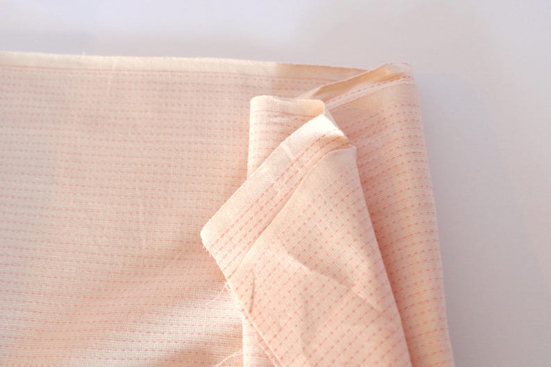 May include: Close-up of a folded piece of peach-colored fabric. The fabric has a textured, dotted pattern and visible stitching. The edges are neatly finished, suggesting a textile or craft material.