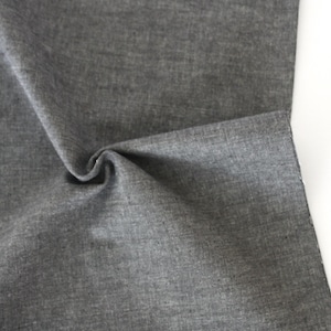 May include: Close-up of a folded piece of gray fabric. The fabric has a textured appearance and a slight sheen. The folds create shadows and depth, highlighting the material's texture and drape. The fabric is against a white background.