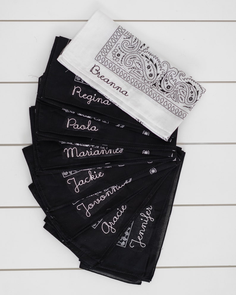 May include: A stack of black bandanas with white embroidered names, including Breanna, Regina, Paola, Marianna, Jackie,  Jovannie, Gracie, and Jennifer. The bandanas are arranged in a fan shape on a white wooden surface.