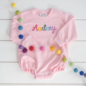 Puede incluir: Un body rosa claro para bebé, con mangas largas y el nombre "Audrey" bordado en colores arcoíris. Una guirnalda de bolas de fieltro coloridas está drapeada sobre el body. Tiene cuello redondo y cierres de presión.