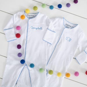May include: Two white baby onesies with blue trim and embroidered names. The onesies are laid out on a white surface with a colorful garland of felt balls.