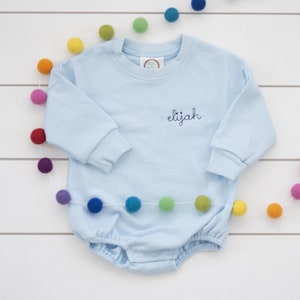 Könnte beinhalten: Hellblauer Baby-Strampler mit langen Ärmeln und Rundhalsausschnitt. Der Strampler hat den Namen "Elijah" in dunkelblauer Farbe auf der Vorderseite gestickt.