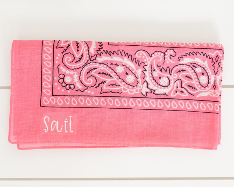 May include: A pink bandana with a white paisley pattern and black border. The bandana has the name "Savil" embroidered in white on the bottom.