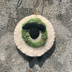 May include: A white felt ornament shaped like a sheep with a green wreath around its head. The wreath is decorated with red, yellow, and blue beads. The sheep has a black face.