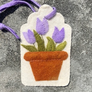 May include: A white felt tag with a purple flower design. The flower is in a brown pot and has three purple flowers with green leaves.