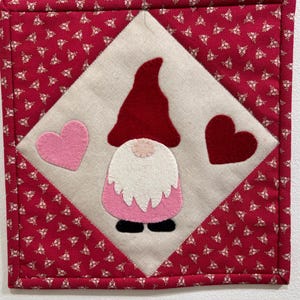 May include: A red and white gnome with a pink skirt and a pink heart on a white background. The gnome is surrounded by a red fabric with a white floral pattern.