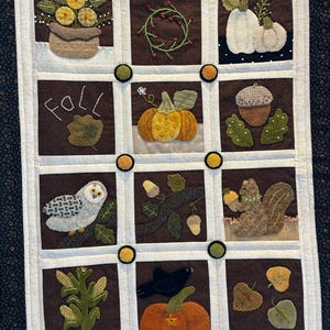 May include: A brown and white quilted wall hanging with a fall theme. The squares feature images of pumpkins, acorns, a squirrel, an owl, a crow, corn, and a wreath. The word "Fall" is stitched in the center of the quilt.