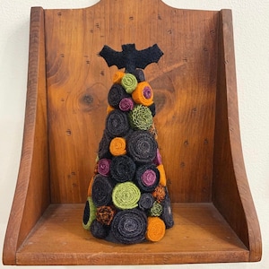May include: A handmade Halloween tree decoration. The tree is cone-shaped and covered in rolled fabric in shades of black, orange, green, and purple. A black felt bat is perched on top. The tree sits on a wooden shelf.