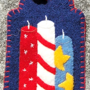 May include: A felt ornament shaped like a tag with three candles. The candles are red, white, and blue with white stars and red stripes. The background is blue.