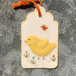 May include: A felt ornament shaped like a tag with a yellow chick, green grass, and a butterfly. The chick is standing on two legs and has a small beak. The butterfly is orange and has its wings spread out. The tag is white with a yellow border.