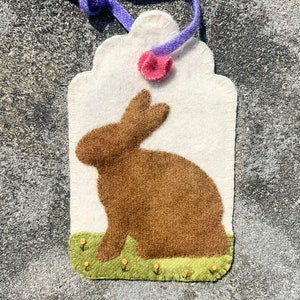 May include: A handmade Easter tag featuring a brown felt bunny on a white background. The tag has a green felt base with small yellow flowers, a purple ribbon, and a pink felt flower. The tag is designed for Easter.