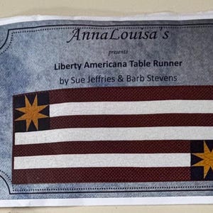 May include: A rectangular table runner with a patriotic design. The runner features alternating burgundy and white stripes, with a dark blue square in each corner containing a gold starburst. The text "AnnaLouisa's presents Liberty Americana Table Runner" is printed on the top.
