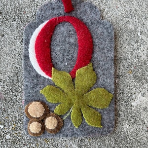 May include: A gray felt ornament with a red felt letter O, a green felt leaf, and three brown felt chestnuts.