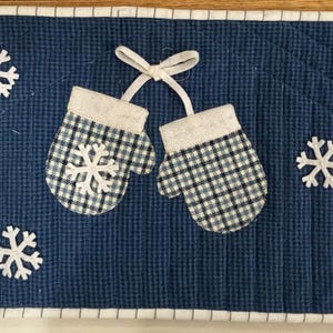 May include: A blue quilted table runner with a white border. The runner features appliqued mittens in plaid patterns, each with a white snowflake and a white cuff. White snowflakes are scattered across the runner.