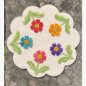 May include: A white felt round rug with a scalloped edge, featuring five colorful felt flowers with green stems and leaves. The flowers are red, orange, blue, purple, and pink.