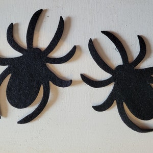 May include: Two black felt spider cutouts. The spiders have eight legs and a rounded body.