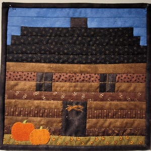 May include: A quilted wall hanging featuring a brown and black cabin with a blue sky above. Two orange pumpkins are in the foreground.