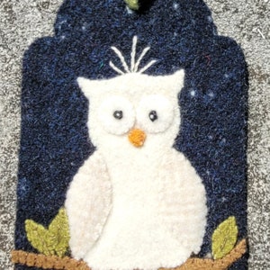 May include: A white felt owl with black eyes and an orange beak perched on a brown branch with green leaves against a dark blue felt background.