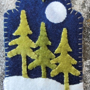 May include: A felt Christmas ornament with a blue background, a white moon, and three green trees. The bottom of the ornament is white, representing snow.