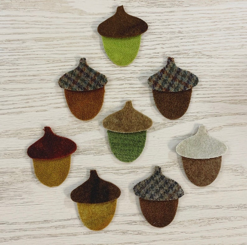 May include: A collection of felt acorn shapes in various colors. The acorn caps are brown, green, and patterned, while the bodies are shades of brown, green, and tan. The acorns are arranged on a light-colored wooden surface.