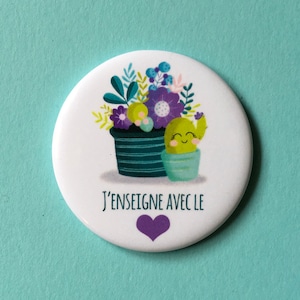 Peut inclure: Un bouton rond blanc avec un motif floral et un cactus. L'illustration présente un cactus souriant dans un pot turquoise à côté d'un pot de fleurs violettes et bleues. Le texte "J'ENSEIGNE AVEC LE" est imprimé sur le bouton, avec un cœur violet en dessous.
