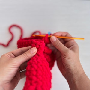 May include: A person's hands crocheting a red knitted item with a crochet hook. The yarn is red and the hook is orange.