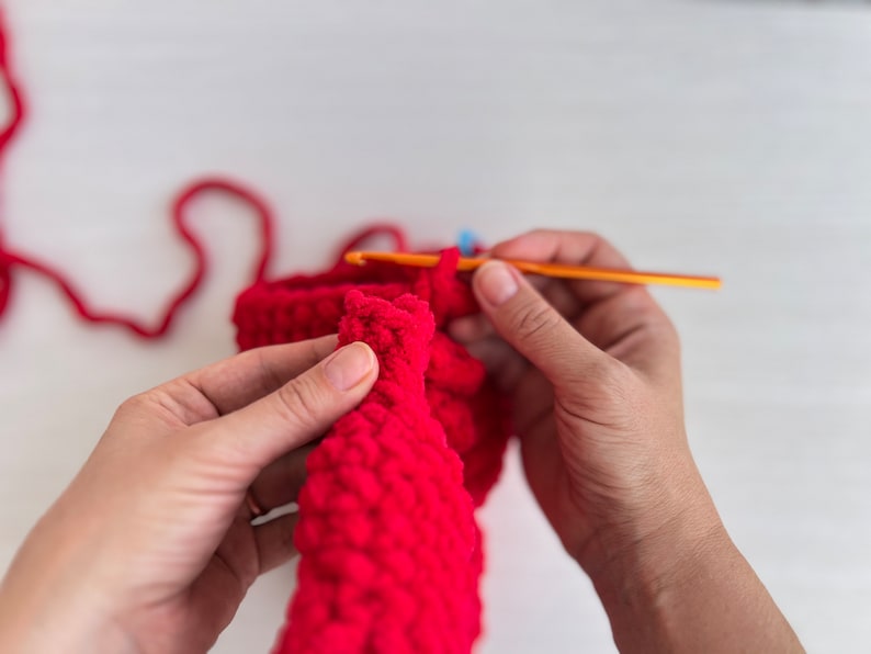 May include: A person's hands crocheting a red knitted item with a crochet hook. The yarn is red and the hook is orange.