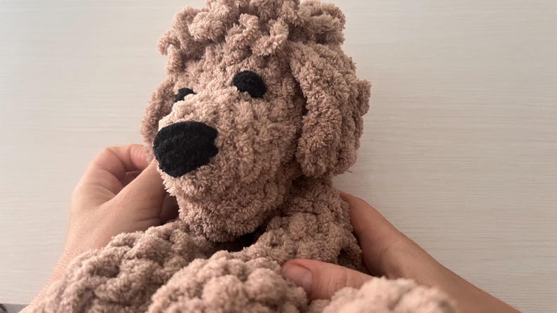 Pode incluir: Um c&atilde;o de pel&uacute;cia feito &agrave; m&atilde;o, em tons de castanho. O c&atilde;o tem uma apar&ecirc;ncia fofa e texturizada com um nariz e olhos pretos. O brinquedo &eacute; segurado por duas m&atilde;os, mostrando seu design macio e aconchegante. Um item charmoso e artesanal.