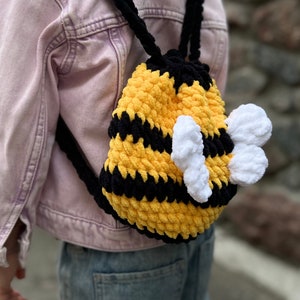 May include: A yellow and black crocheted bee backpack with white wings. The backpack is being worn by a child.