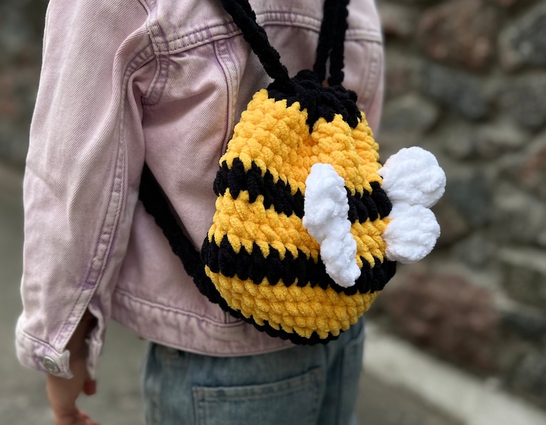 May include: A yellow and black crocheted bee backpack with white wings. The backpack is being worn by a child.