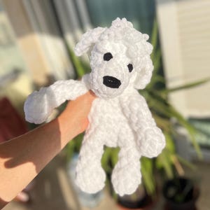 Pode incluir: Um brinquedo de cachorro de croch&ecirc; branco feito &agrave; m&atilde;o com olhos pretos e um nariz preto. O brinquedo de pel&uacute;cia tem uma textura fofa e &eacute; segurado em frente a um fundo desfocado de vegeta&ccedil;&atilde;o e luz solar.
