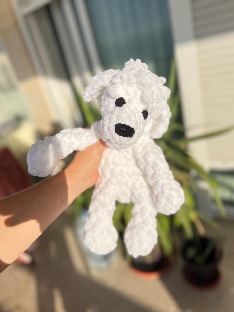 Pode incluir: Um brinquedo de cachorro de croch&ecirc; branco feito &agrave; m&atilde;o com olhos pretos e um nariz preto. O brinquedo de pel&uacute;cia tem uma textura fofa e &eacute; segurado em frente a um fundo desfocado de vegeta&ccedil;&atilde;o e luz solar.