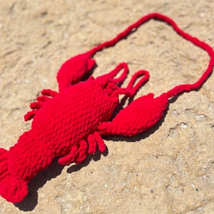 May include: A red crocheted lobster toy with a long tail. The toy is lying on a sandy surface.