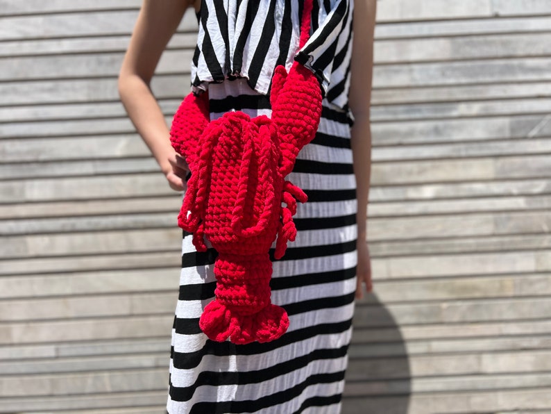 May include: A red crocheted lobster bag worn as a crossbody bag by a person wearing a black and white striped dress.
