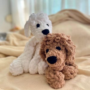 Pode incluir: Dois bichos de pel&uacute;cia de cachorro de croch&ecirc; feitos &agrave; m&atilde;o. Um &eacute; branco, o outro &eacute; marrom. Ambos t&ecirc;m olhos e nariz pretos. Os c&atilde;es est&atilde;o sentados em um cobertor bege macio.