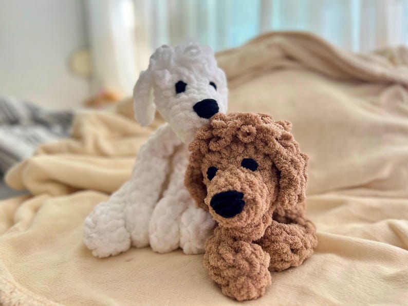 Pode incluir: Dois bichos de pel&uacute;cia de cachorro de croch&ecirc; feitos &agrave; m&atilde;o. Um &eacute; branco, o outro &eacute; marrom. Ambos t&ecirc;m olhos e nariz pretos. Os c&atilde;es est&atilde;o sentados em um cobertor bege macio.